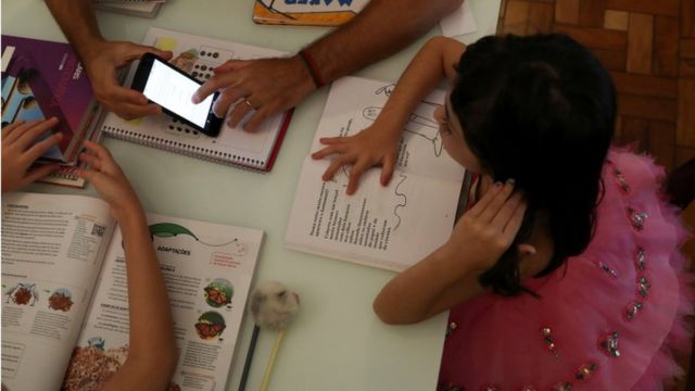 Em foto tirada de cima, duas crianças com seus livros didáticos e um adulto mostrando algo no celular