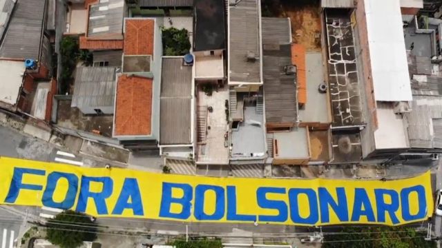 Faixa amarela colocada na rua e vista do alto diz 'fora bolsonaro' em azul; ao lado, telhados de casas