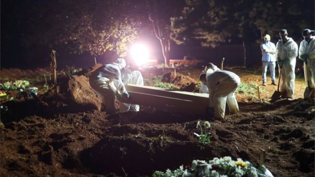 Coveiros vestindo roupas de proteção carregam o caixão de um homem de 32 anos que morreu por covid enquanto holofotes iluminam os túmulos durante os enterros noturnos no cemitério de Vila Formosa em São Paulo, Brasil, 30 de março de 2021