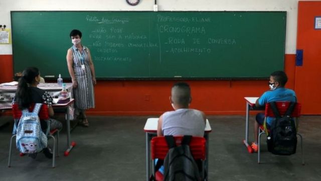 Alunos e professora de máscara e mantendo distanciamento social entre as carteiras na sala de aula