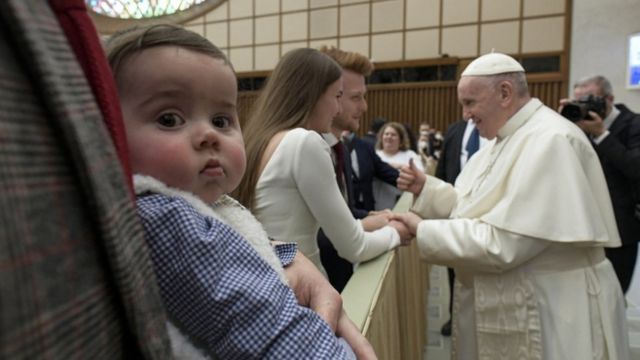 Bebê olha para a câmera, com papa conversando com casal em segundo plano